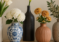Five ceramic vases in white matte, blue-and-white glaze, black, terracotta and speckled gray holding cream and blush artificial flowers on a wood console